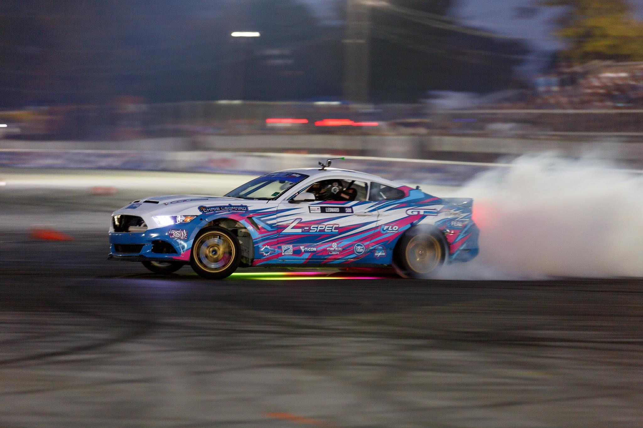 Rennspec-designed blue, pink and white motorsports wrap on a Mustang drifting at speed